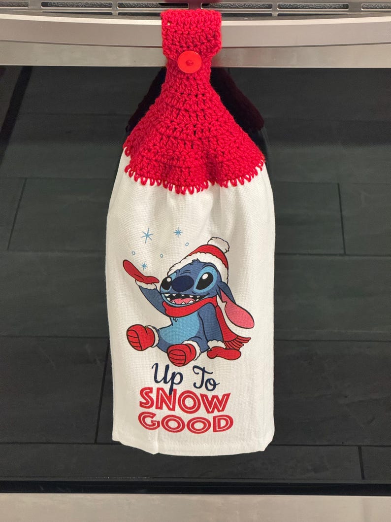 Puede incluir: Un pa&ntilde;o de cocina blanco con una parte superior y un bot&oacute;n de ganchillo rojos. La toalla presenta un personaje de dibujos animados con gorro y bufanda de invierno, con el texto "Up To SNOW GOOD" en rojo.