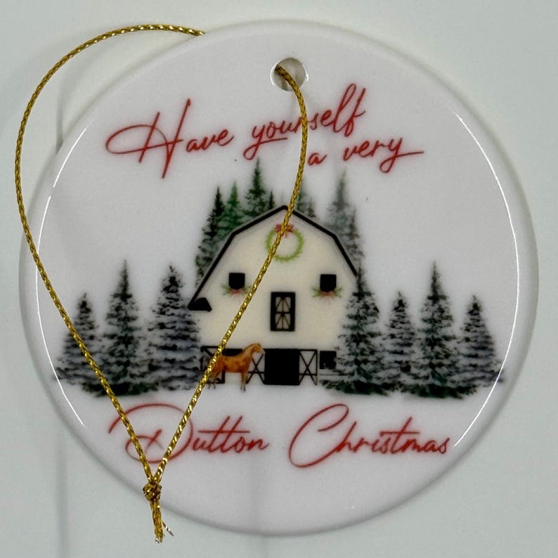 May include: White ceramic Christmas ornament with a gold string. The ornament features a barn scene with a horse and pine trees. Text reads "Have yourself a very Dutton Christmas" in red script.