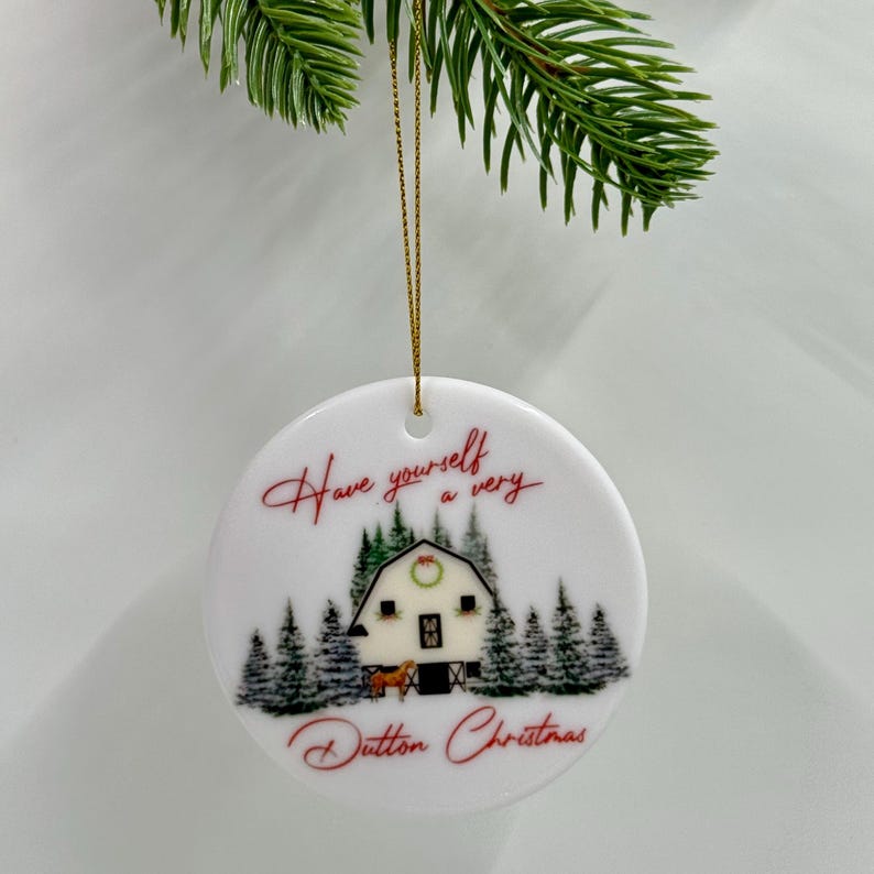 May include: White ceramic Christmas ornament with a gold string. The ornament features a winter scene with a barn, trees, and the text "Have yourself a very Dutton Christmas" in red script.