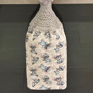 May include: A gray crocheted kitchen towel holder with a button closure. The towel features a white background with a repeating pattern of the character Stitch from the movie Lilo & Stitch in blue, pink, and gray.