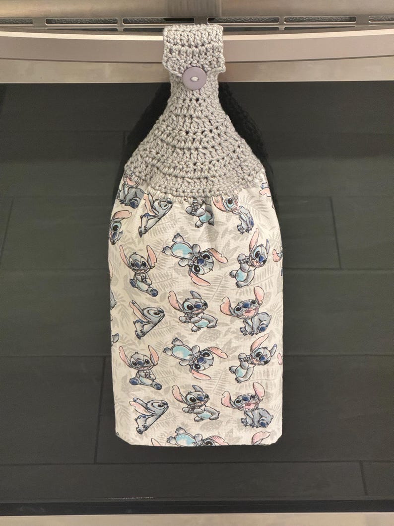 May include: A gray crocheted kitchen towel holder with a button closure. The towel features a white background with a repeating pattern of the character Stitch from the movie Lilo & Stitch in blue, pink, and gray.