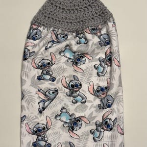 May include: A hanging kitchen towel featuring a gray crocheted top with a button closure. The towel's body is white with a repeating pattern of the cartoon character Stitch in various poses, set against a backdrop of gray leaves.