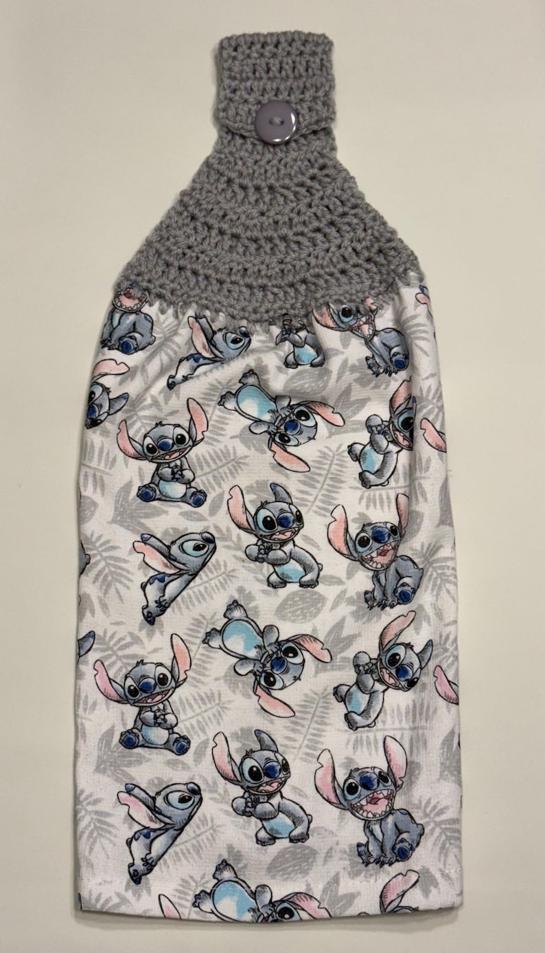 May include: A hanging kitchen towel featuring a gray crocheted top with a button closure. The towel's body is white with a repeating pattern of the cartoon character Stitch in various poses, set against a backdrop of gray leaves.