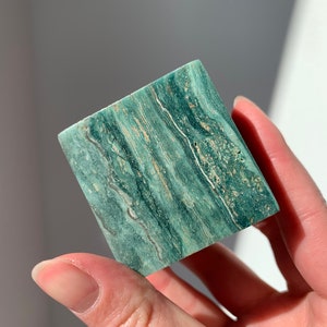 May include: A square-shaped stone with a teal and tan striped pattern. The stone is held in a hand and appears to be a decorative item or a piece of natural art. The stone's surface has a polished appearance.