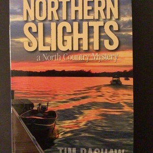 May include: A book cover with the title "NORTHERN SLIGHTS" in large, beige letters. Below, it reads "a North Country Mystery." The cover art depicts a sunset over a lake with boats and a dock. The author's name, "TIM BASHAW," is at the bottom.