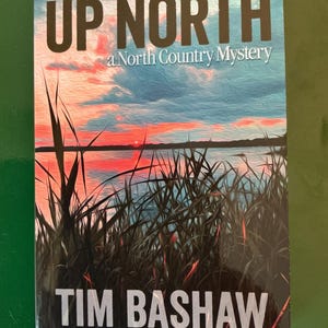 May include: A book cover with the title "UP NORTH" in large, dark letters above the subtitle "a North Country Mystery." The cover features a scenic painting of a lake at sunset, with tall grass in the foreground and the author's name, Tim Bashaw, at the bottom.