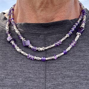 May include: A layered necklace featuring amethyst beads and silver-toned metal accents. The necklace has a mix of small, round beads and larger, square-cut amethyst stones. The design is intricate and the color palette is primarily purple and silver.