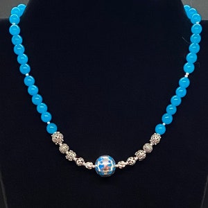 Puede incluir: Un collar con cuentas turquesas brillantes con detalles plateados. El collar tiene una cuenta central grande y estampada y cuentas decorativas más pequeñas. El collar se muestra sobre un fondo negro.