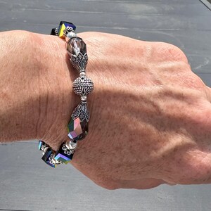 May include: A beaded bracelet featuring faceted, iridescent crystals and silver-toned metal beads. The bracelet includes a mix of cube and teardrop-shaped crystals, along with decorative round and cone-shaped metal beads. The bracelet is worn on a wrist.