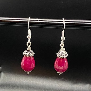 May include: A pair of drop earrings featuring faceted, teardrop-shaped, deep pink gemstones. The earrings have silver-coloured metal accents and hooks. The gemstones are suspended from ornate silver-toned caps.