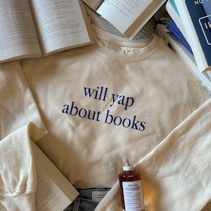 May include: A cream colored sweatshirt with the text "will yap about books" printed in blue on the front. The sweatshirt is laying on a table with a book and a bottle of perfume.
