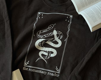 Puede incluir: Sudadera negra con un gráfico blanco de una serpiente en un marco con el texto "the romantasy reader".