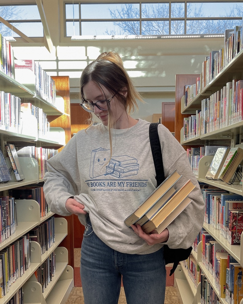 以下が含まれることがあります： 「Books are my friends」と書かれたグレーのトレーナーを着た人が、本を手に持っている。トレーナーには、笑顔の顔をした本の積み重ねの漫画イラストが描かれている。その人は図書館にいる。