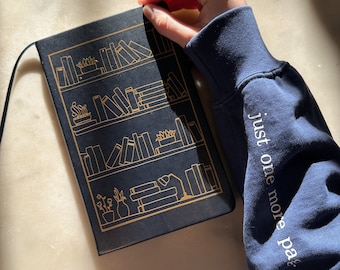 May include: A black notebook with a gold design of a bookshelf filled with books. The notebook has the text "just one more page..." printed on the cover.