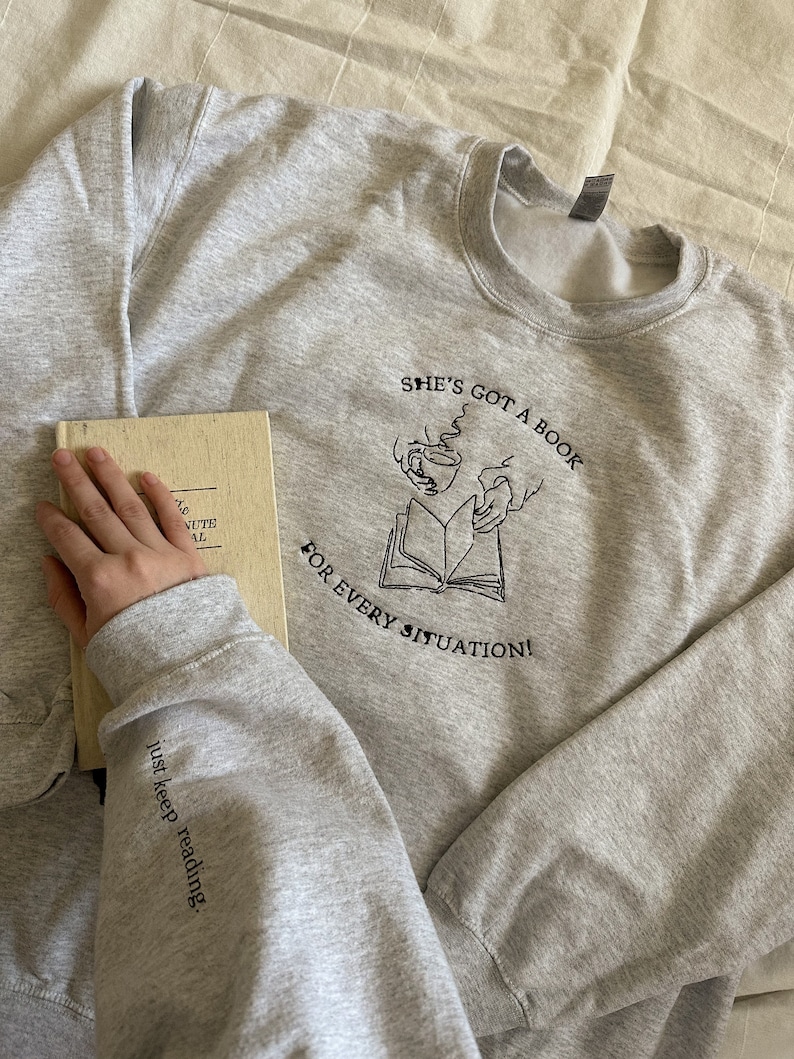 May include: Gray sweatshirt with a black graphic of a book and two hands holding cups. The text "She's got a book for every situation!" is printed on the sweatshirt. The text "just keep reading." is printed on the sleeve.