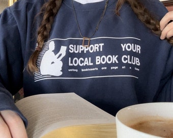 Puede incluir: Una persona lleva una sudadera azul marino con el texto "Support Your Local Book Club" y una silueta blanca de una persona leyendo un libro. El texto debajo del gráfico dice "Uniting booktroverts one page at a time".
