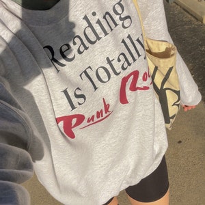 May include: A gray sweatshirt with the text "Reading is Totally Punk Rock" printed on the front. The sweatshirt is being worn by a person wearing black shorts and gray sneakers.