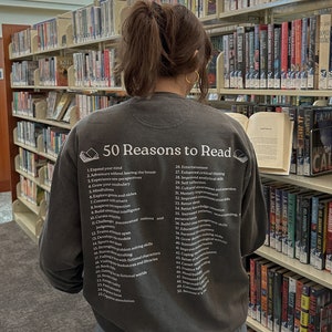 May include: A dark gray sweatshirt with the text "50 Reasons to Read" printed on the back. The text is in white and is surrounded by a small book icon. The list of reasons is in white text and includes items such as "Expand your mind", "Adventure without leaving the house", "Experience new perspectives", and "Grow your vocabulary".