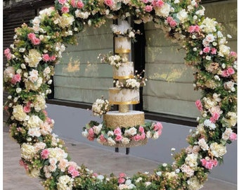 May include: A white wedding cake with gold accents is suspended from a floral archway made of white and pink flowers. The archway is made of greenery and flowers.