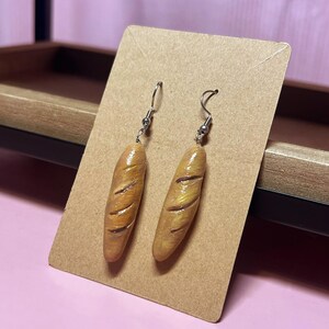 Könnte beinhalten: Ein Paar ausgefallene Ohrringe in Form von Miniatur-Baguettes. Die Ohrringe sind hellbraun mit dunkleren braunen Linien, die die Brotkruste nachahmen. Sie hängen an silberfarbenen Fischhaken-Ohrdrähten.