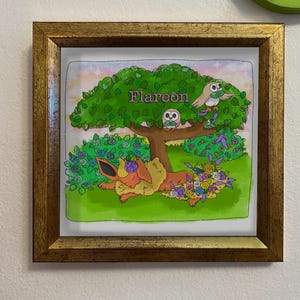 May include: A framed artwork featuring a cartoon Flareon character resting under a tree with the name "Flareon" above. The artwork includes owls, flowers, and a green and gold color scheme.