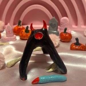 May include: A black, arch-shaped clay figurine with two red horns and a central red circular accent. The figurine is positioned among small orange pumpkins, gray and pink tombstones, and a light blue, red-tipped object on a satin fabric.