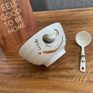 May include: White ceramic bowl with a floral design and gold accents. The bowl is sitting on a wooden table next to a white ceramic spoon with a floral design.
