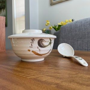 May include: White ceramic bowl with a lid and a floral design. The bowl has a gold accent and Japanese writing. A white plastic spoon is next to the bowl.