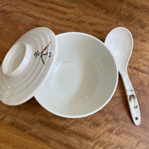 May include: A white ceramic bowl with a speckled finish and a matching lid. The lid has a brown design with Japanese characters. A white ceramic spoon with a brown design is also included.
