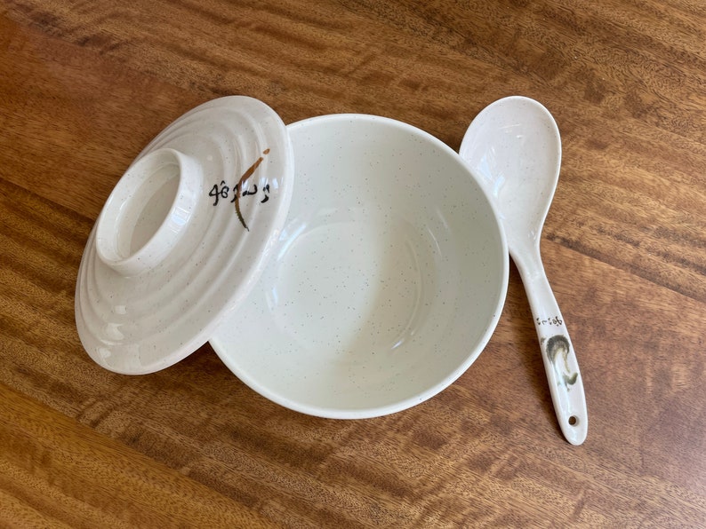 May include: A white ceramic bowl with a speckled finish and a matching lid. The lid has a brown design with Japanese characters. A white ceramic spoon with a brown design is also included.