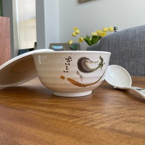 May include: White ceramic bowl with a lid and a matching spoon. The bowl has a gold and green design with Japanese characters.