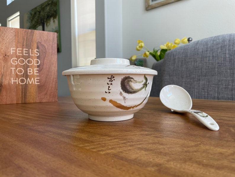 May include: White ceramic bowl with a lid and a floral design. The bowl has a gold accent and Japanese writing. A white plastic spoon is next to the bowl.