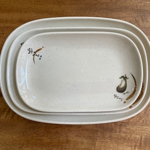 Puede incluir: Tres platos de servir ovalados de cerámica blanca con un acabado moteado. El plato más grande tiene una ilustración de berenjena marrón y verde y texto japonés. Los platos más pequeños están anidados dentro del más grande.