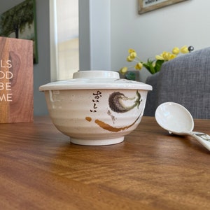 May include: A white ceramic bowl with a lid and a floral design. The bowl has Japanese writing on the side. A white plastic serving spoon is next to the bowl.