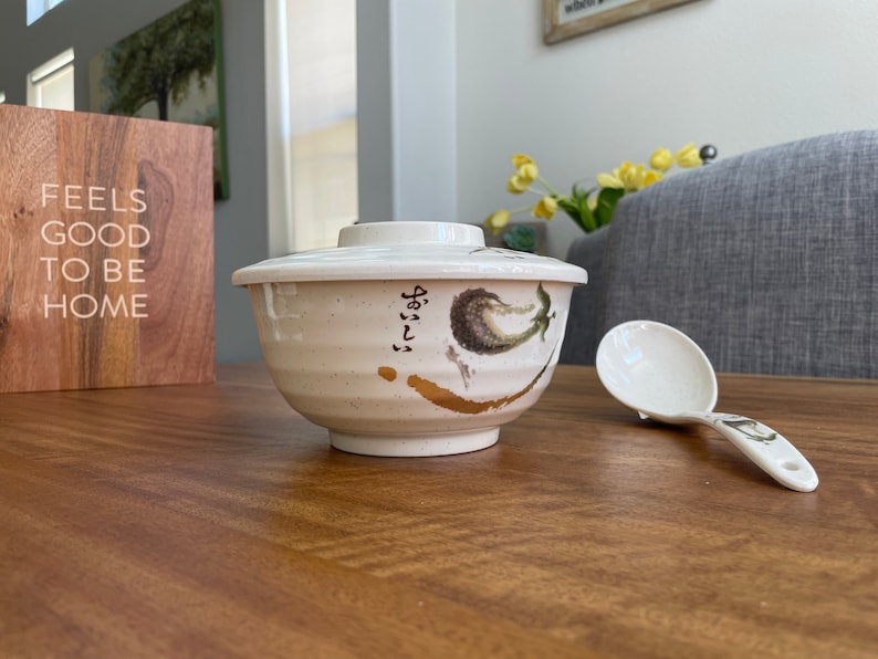May include: A white ceramic bowl with a lid and a floral design. The bowl has Japanese writing on the side. A white plastic serving spoon is next to the bowl.