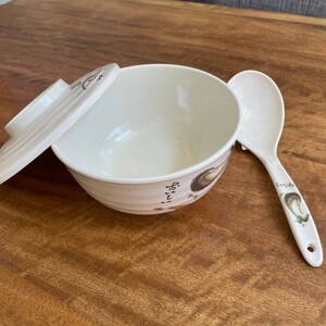 May include: A white ceramic bowl with a lid and a matching white spoon. The bowl has a speckled design and a floral pattern on the side. The spoon has a floral design on the handle.