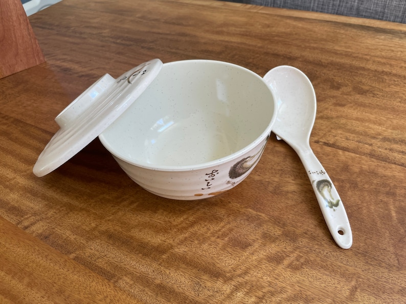 May include: A white ceramic bowl with a lid and a matching white spoon. The bowl has a speckled design and a floral pattern on the side. The spoon has a floral design on the handle.