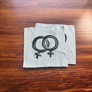 May include: Two square light grey fabric patches featuring a black screen-printed design of two interlocking female gender symbols. The patches have raw, unfinished edges and are made of a woven textile material with a visible diagonal twill pattern.