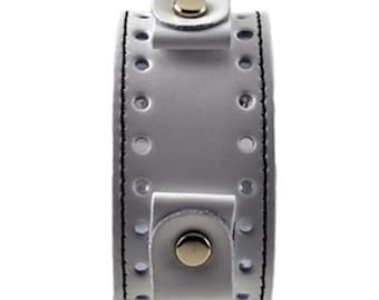 May include: A light gray leather watch cuff with a snap closure and gold-colored metal accents. The cuff features decorative stitching and evenly spaced holes along the edges.