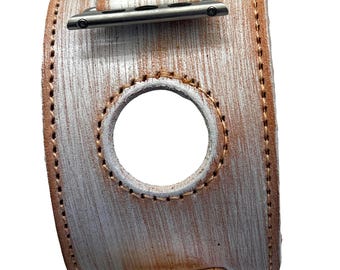 May include: A distressed leather watch band with a white and brown wood grain pattern. The band features a large circular opening and is secured with silver-tone metal hardware. The edges are stitched with brown thread.