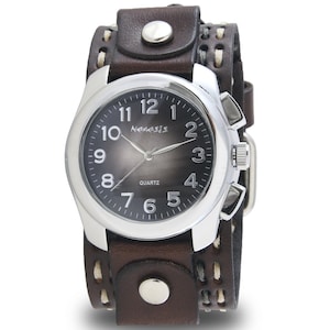 Puede incluir: Un reloj de cuero marrón con esfera plateada y correa ancha. La esfera es gris oscuro con números blancos y la palabra "Nemesis". La correa tiene costuras blancas y tachuelas plateadas.