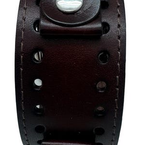 May include: A wide dark brown leather cuff bracelet featuring two vertical rows of circular cutouts along the center. The cuff is secured with two silver-toned metal snap buttons at the top and bottom, with visible stitching along the edges.