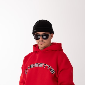 Könnte beinhalten: Ein roter Kapuzenpullover mit dem Wort "CASSETTE" in Schwarz-Weiß über der Brust. Die Person trägt eine schwarze Mütze und eine Sonnenbrille. Der Kapuzenpullover hat eine Vordertasche.