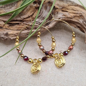 Könnte beinhalten: Goldene Creolen mit einem Spiral-Anhänger. Die Creolen sind mit kleinen goldenen Perlen und roten und gelben Glasperlen verziert.