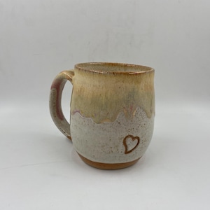 May include: A ceramic mug with a rounded body and a curved handle. The upper portion features a smooth, tan-colored glaze that drips over a speckled, off-white base. A small, hand-drawn brown heart outline is centered on the lower front section.