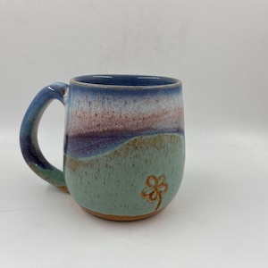 May include: A ceramic mug featuring a speckled glaze with a colour gradient transitioning from light green at the base to purple and blue at the rim. The mug has a curved handle and a small, hand-drawn flower outline etched into the lower front.