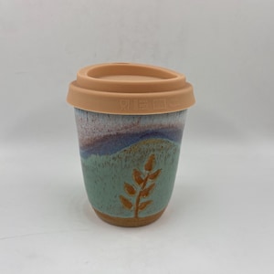 May include: A ceramic travel mug with a light peach silicone lid. The mug features a speckled glaze with a gradient of light green at the base, transitioning to blue and white at the top. A brown leaf design is painted on the front surface.