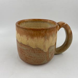 May include: A handcrafted ceramic mug with a rounded body and a large handle. The mug has a colour gradient, shifting from a reddish-brown base to a tan and light brown upper section. The mug is designed for hot drinks.