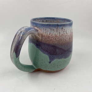 May include: A ceramic mug with a rounded body and a large handle. The mug has a colour gradient, transitioning from blue at the rim, through brown and purple, to a green base. Suitable for hot or cold drinks, the mug is handmade.