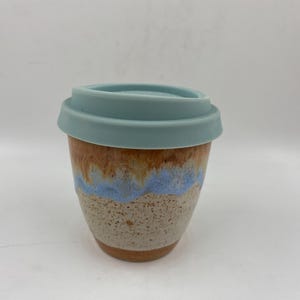 May include: A ceramic travel mug with a light blue silicone lid. The mug features a speckled beige base that transitions into a wavy band of light blue glaze, topped with a smooth, warm brown drip effect near the rim of the vessel.
